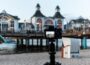 Reisestativ mit Kamera am Strand, im Hintergrund eine Seebrücke.