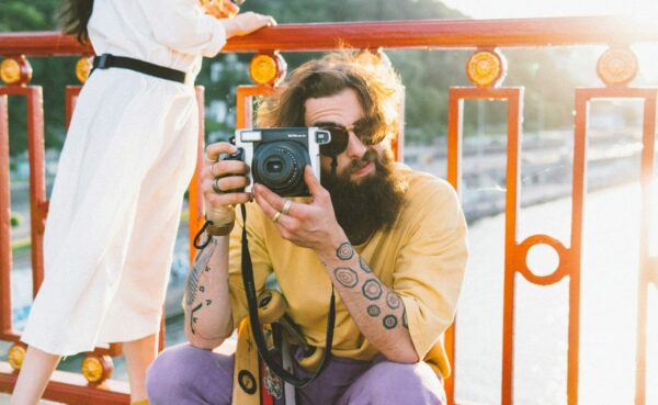 Mann mit Vollbart und Sonnenbrille hält eine Fujifilm Instax Sofortbildkamera, sitzend auf einer Brücke bei Sonnenuntergang.