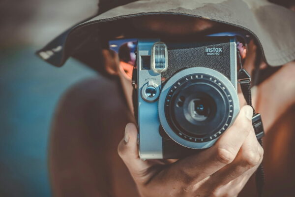 Person mit braunem Fischerhut hält eine schwarze und graue Fujifilm Instax Sofortbildkamera, bereit, ein Foto aufzunehmen.