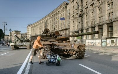 Person schiebt Kinderwagen vorbei an zerstörten Panzern auf einer städtischen Straße, Symbol für Reportagefotografie in Kriegs- und Krisengebieten.